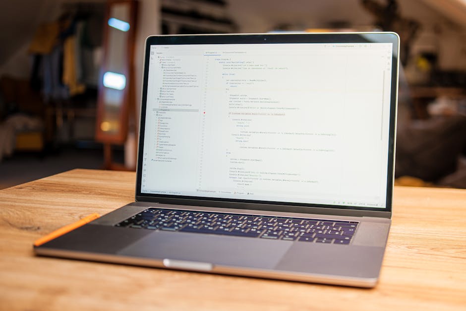 A modern silver laptop placed on a wooden table in a well-lit room, displaying lines of code on the screen. The laptop has a black keyboard with a touchpad below, and an orange pencil rests on the lef