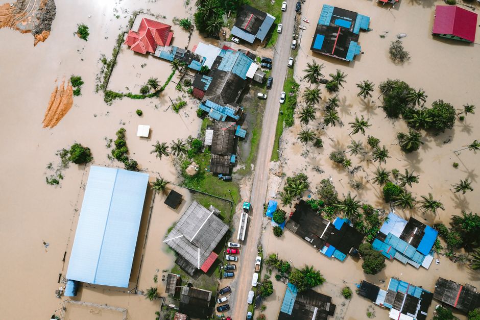 An aerial view of a residential area affected by flooding, showing houses with partially submerged roofs and yards. The floodwaters cover large portions of the ground, with muddy and murky surfaces vi